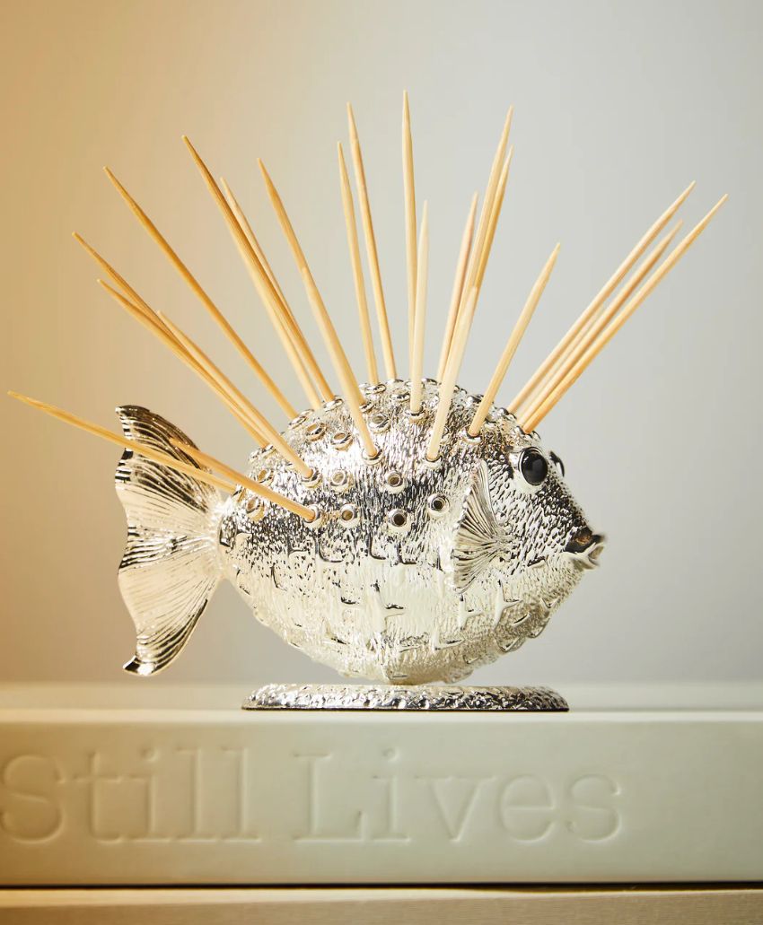 A metallic pufferfish-shaped holder with wooden toothpicks sticking out like spines, placed on top of a closed book with the partially visible title Still Lives.