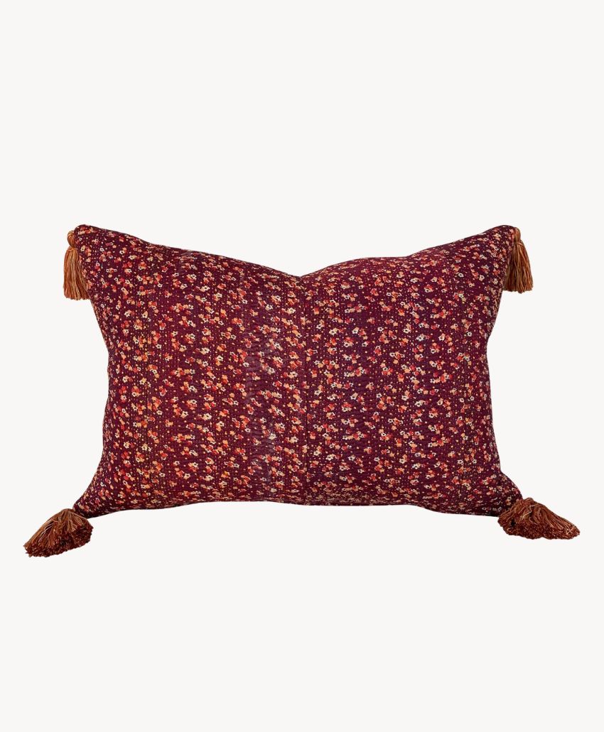 Rectangular pillow with a burgundy background and small floral pattern, featuring four brown tassels on each corner.
