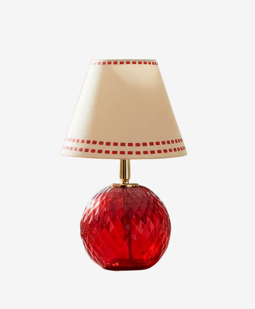 A table lamp with a round, faceted red glass base and a cream-colored lampshade decorated with a border of small red squares.