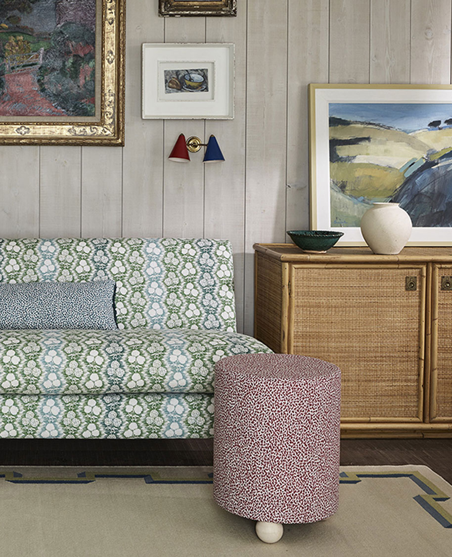 A cozy living room with a green and white patterned sofa, a red patterned round stool, a wooden cabinet with art and ceramics on top, and paintings hanging on wood-paneled walls.