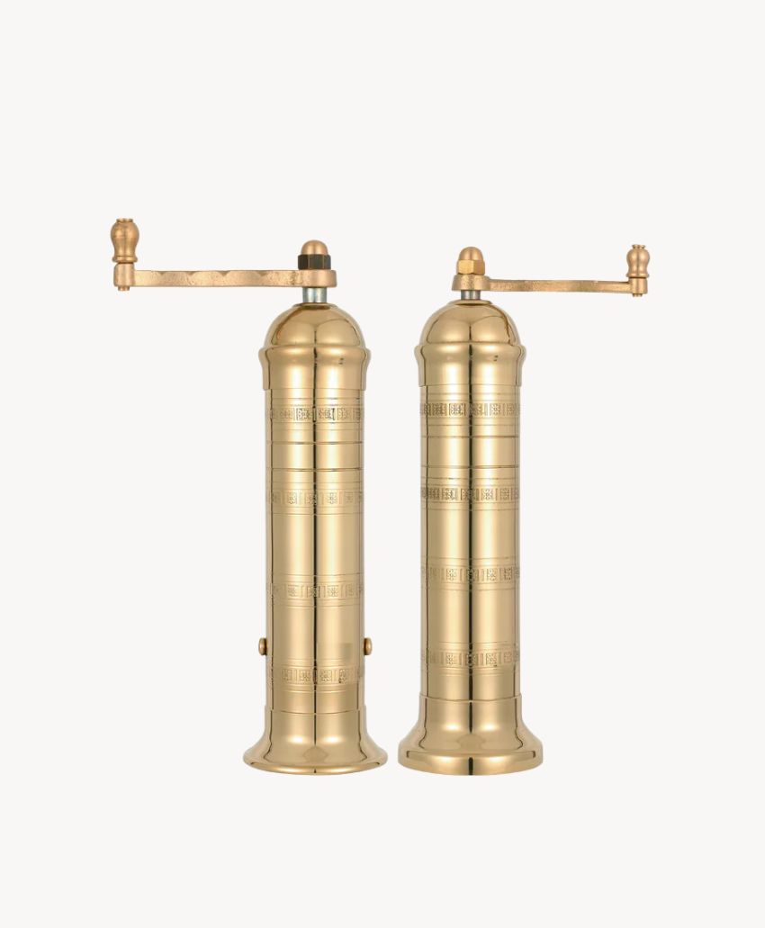 Two tall, cylindrical brass pepper or spice mills with engraved patterns and long horizontal handles, standing side by side on a plain white background.