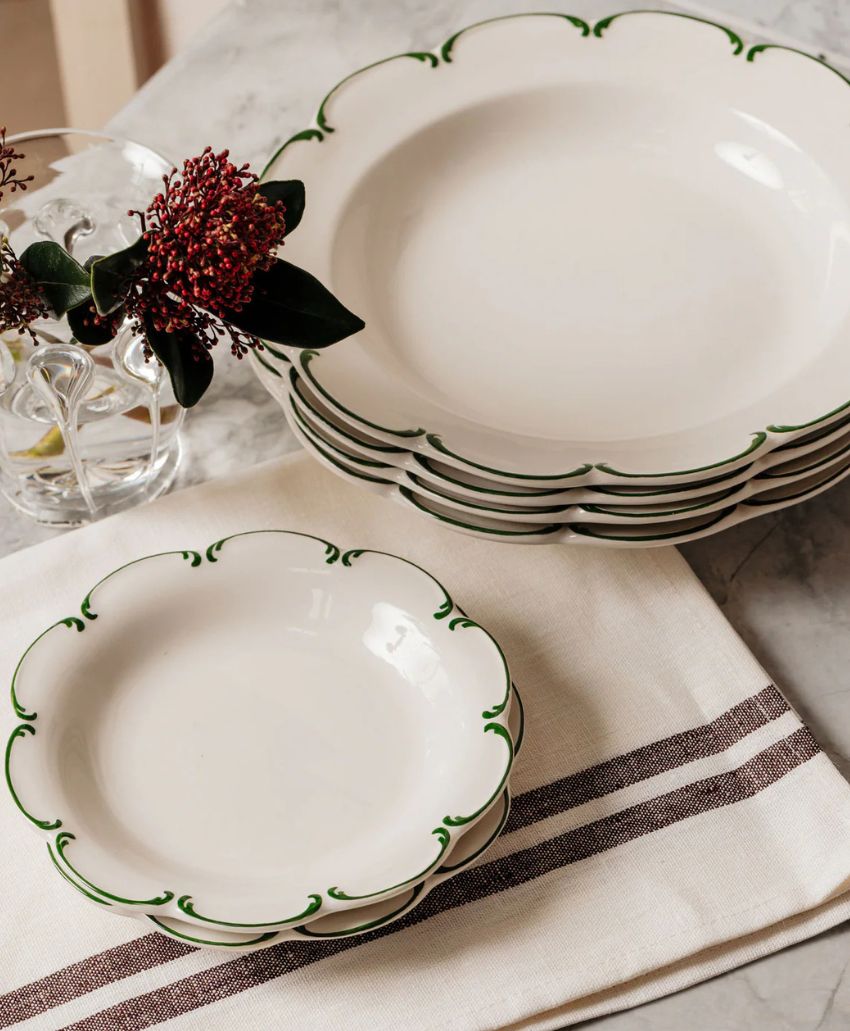 A stack of white scalloped plates with green trim sits on a striped cloth next to a glass vase holding red flowers and greenery on a marble surface.
