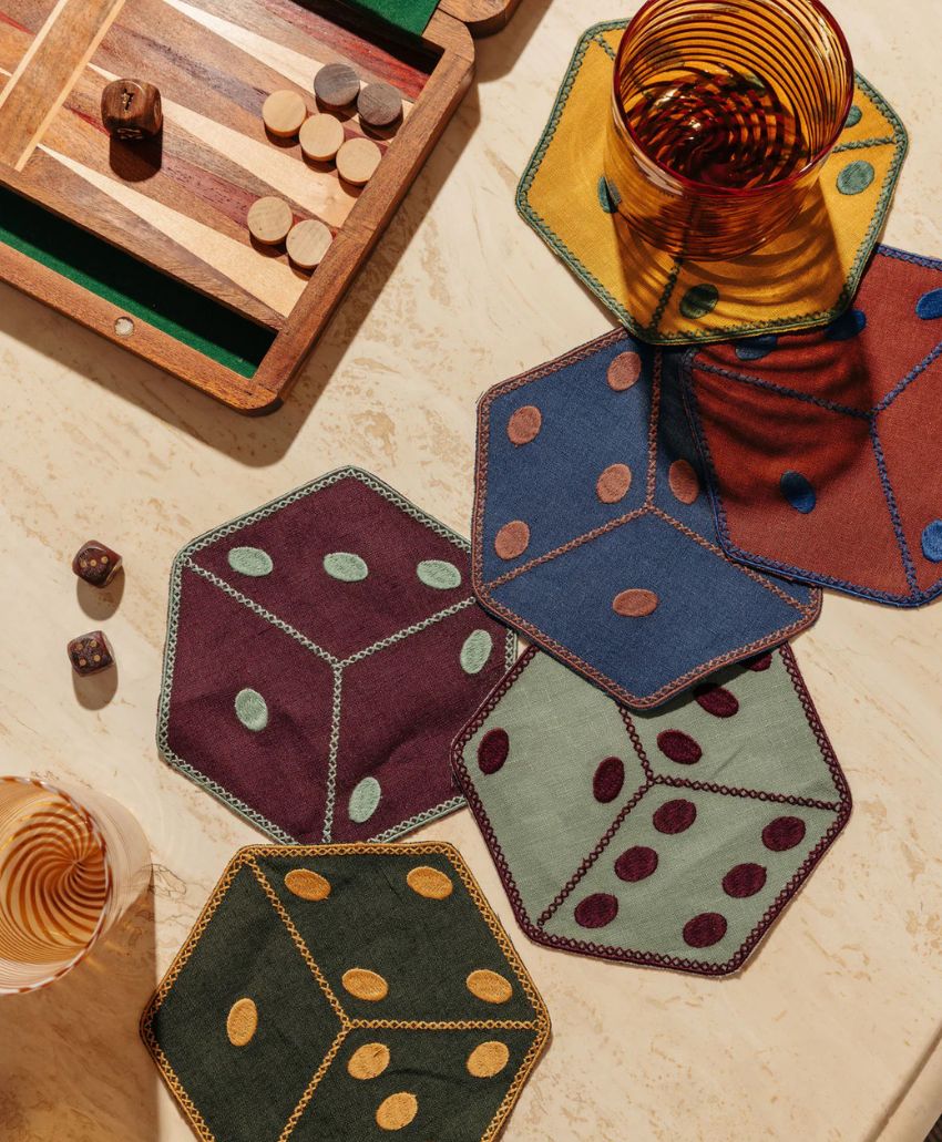 A backgammon board and dice rest on a beige surface beside a striped glass and several colorful, hexagon-shaped coasters designed to look like dice.