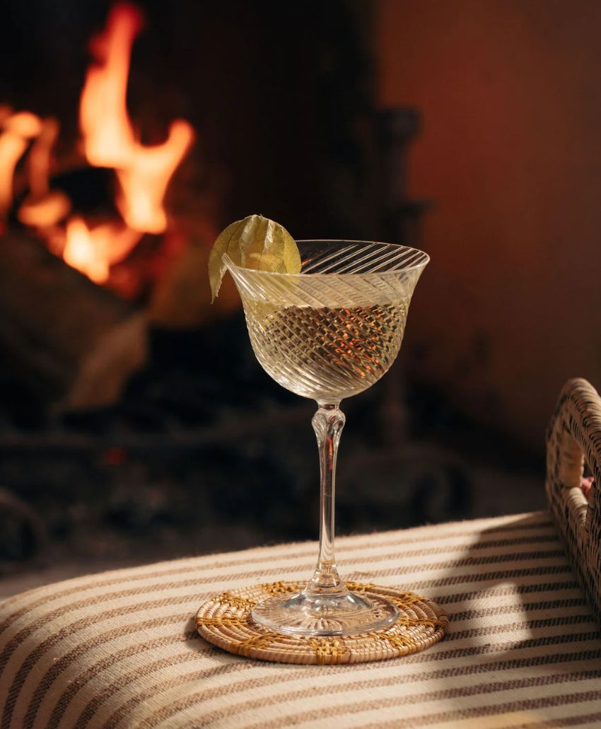 A crystal cocktail glass with a lime garnish sits on a woven coaster atop a striped cushion, with a warm, blurred fireplace burning in the background.