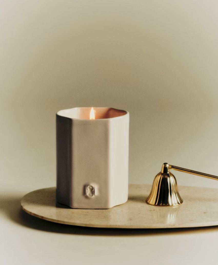 A lit, white, square candle sits on a beige oval tray next to a gold candle snuffer, with a neutral background.