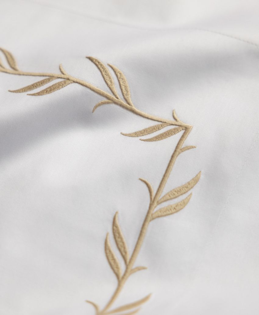 Close-up of white fabric with a decorative beige embroidered leaf and stem pattern forming an elegant corner design.