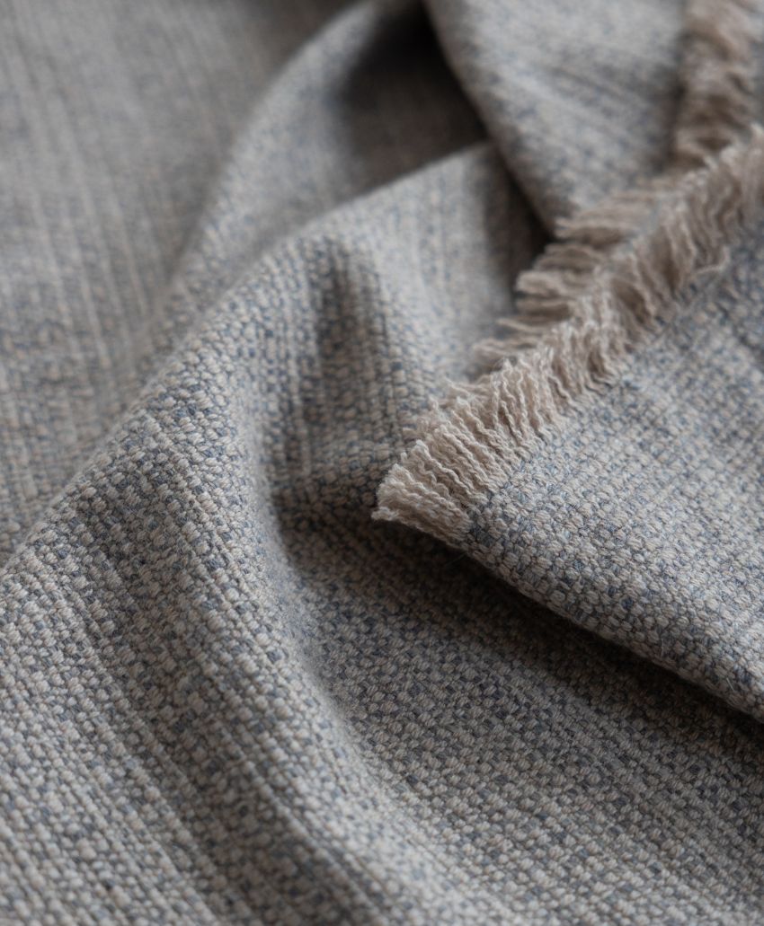 A close-up of a textured, light gray woven fabric with a fringed edge, showing its detailed pattern and soft folds.