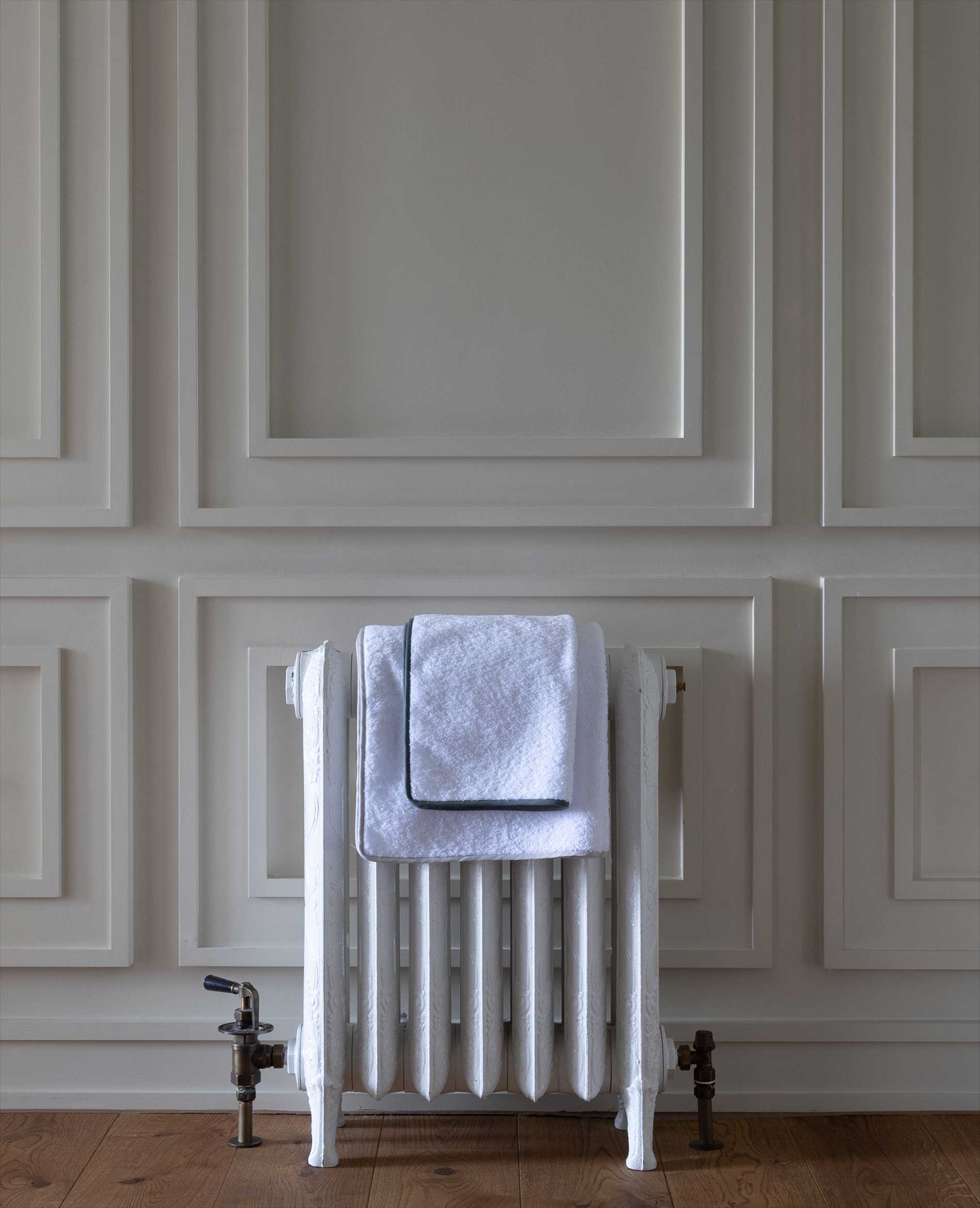 A white towel with a gray border is neatly folded and placed on top of a vintage white radiator, set against a wall with decorative rectangular panel molding and a wooden floor.