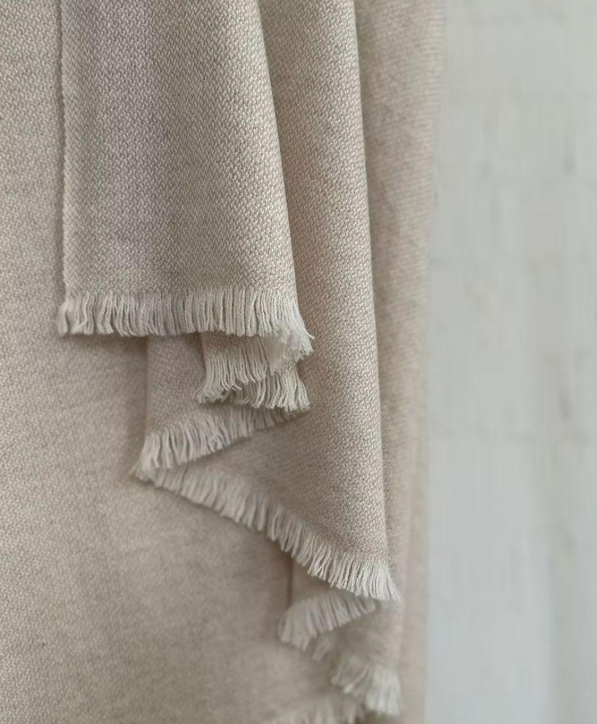 A close-up of a beige woven fabric with fringed edges, draped in soft folds against a light, neutral background.