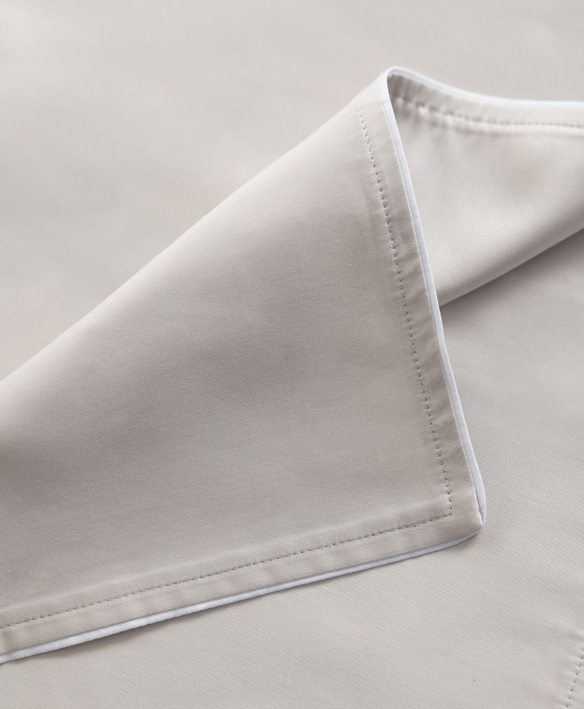 A close-up of a neatly folded light beige fabric napkin with clean, visible stitching along the edges, resting on a matching fabric surface.
