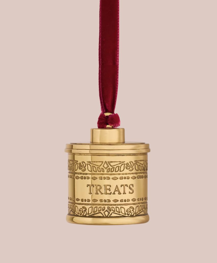 A gold, cylindrical ornament with TREATS engraved on it, decorated with leaf patterns, and hanging from a red velvet ribbon against a light beige background.