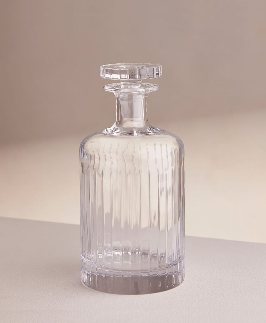 A clear glass decanter with vertical ridges and a matching glass stopper, standing on a smooth, light-colored surface against a soft, neutral background.