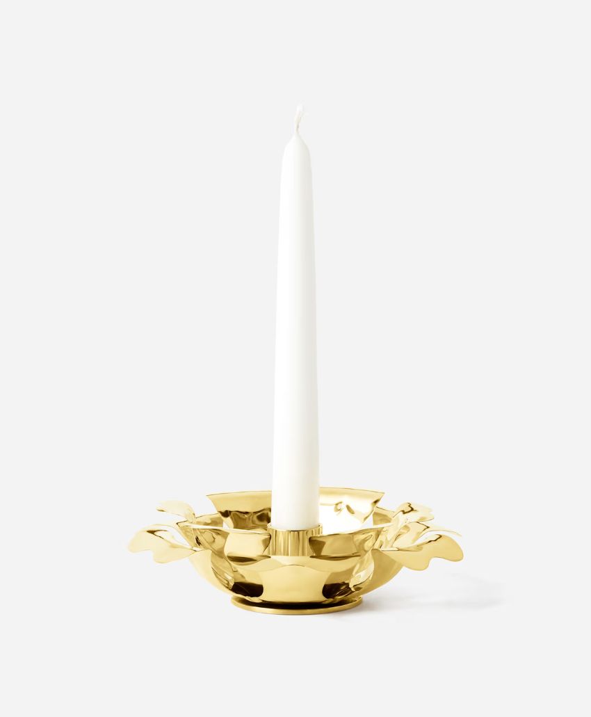 A white taper candle stands upright in a shiny gold candle holder with leaf-like decorative edges, set against a plain white background.