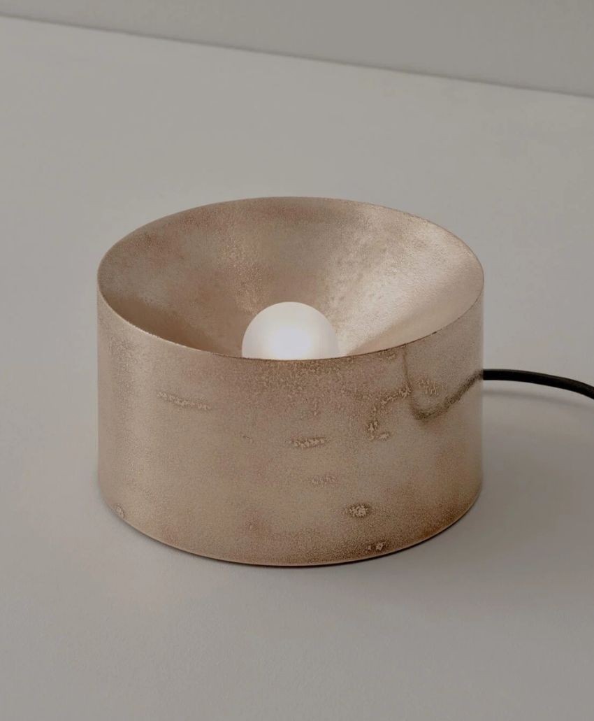 A metallic, cylindrical table lamp with a rough, textured surface and a recessed white bulb in the center, placed on a light-colored surface with a visible black power cord.