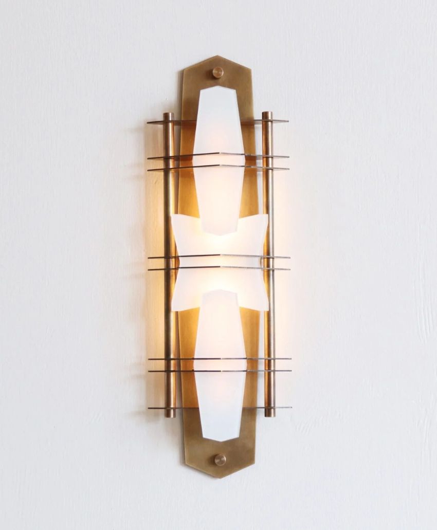 A modern wall sconce with a brass frame, white geometric glass shade, and horizontal metal rods, mounted on a light-colored wall and casting a warm glow.