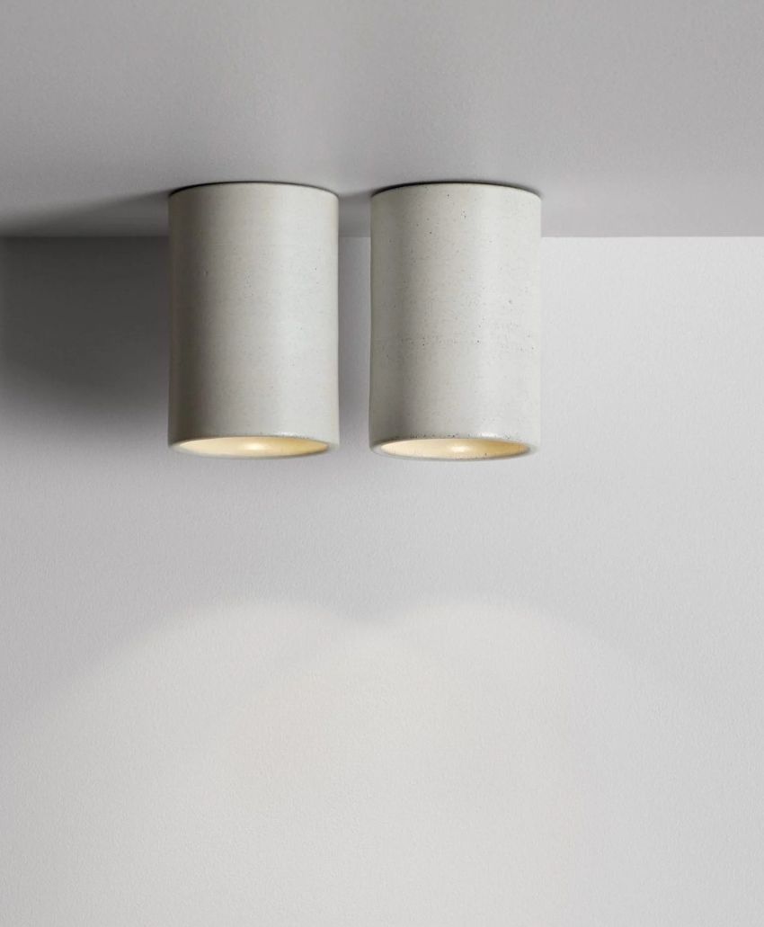 Two cylindrical, modern ceiling light fixtures with a smooth, matte white finish are mounted side by side on a white ceiling, casting soft light onto a plain light gray wall below.