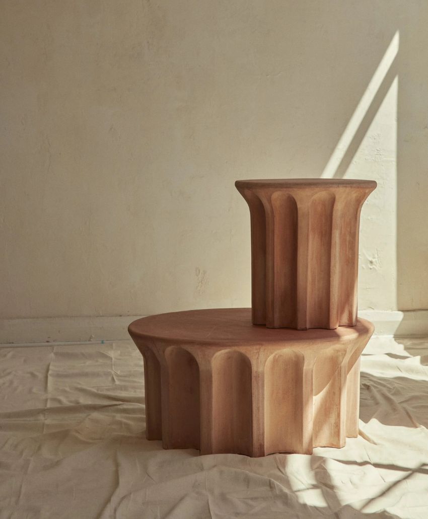 Two sculptural wooden objects with fluted sides, one tall and one short, are stacked on a draped fabric against a plain, sunlit wall. Light and shadows highlight their smooth, geometric shapes.