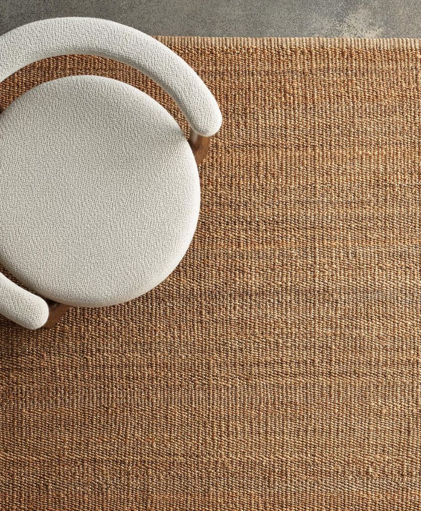 A curved, upholstered white chair sits on a textured, light brown woven rug, viewed from above on a gray concrete floor.