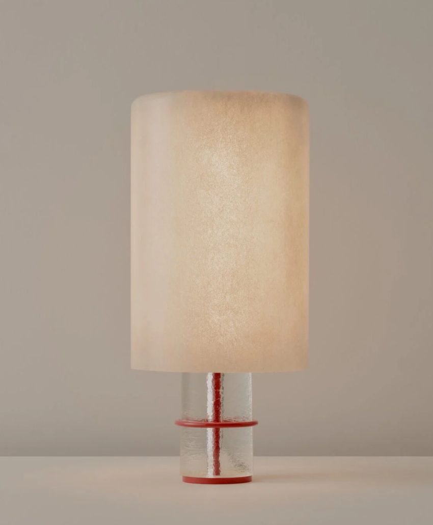 A modern table lamp with a cylindrical, textured beige shade and a silver base with a red accent, placed on a light surface against a plain, neutral background.