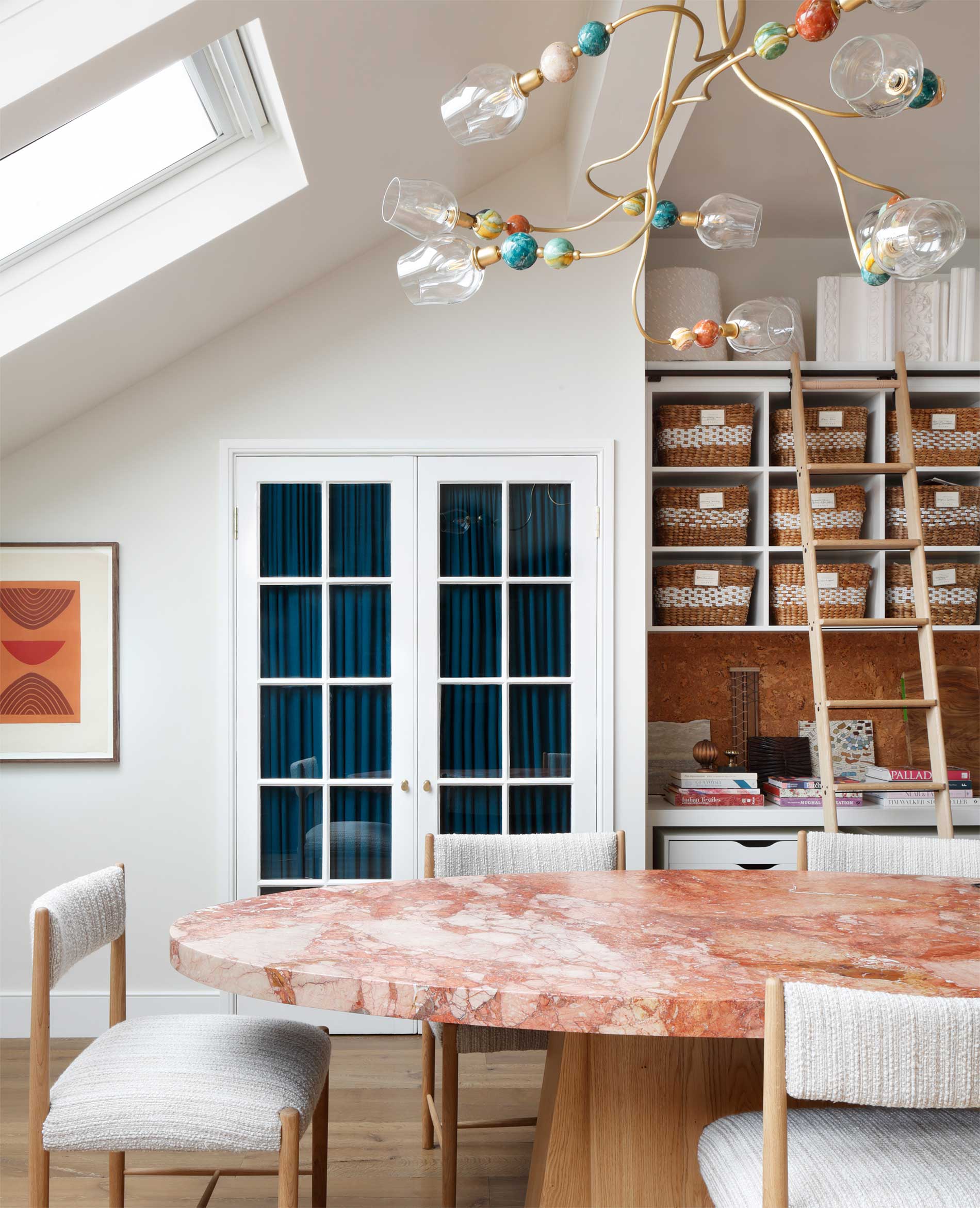 A modern dining area with a pink marble table, upholstered chairs, woven baskets on shelves, a colorful beaded chandelier, and a skylight above. French doors and wall art complete the bright, airy space.