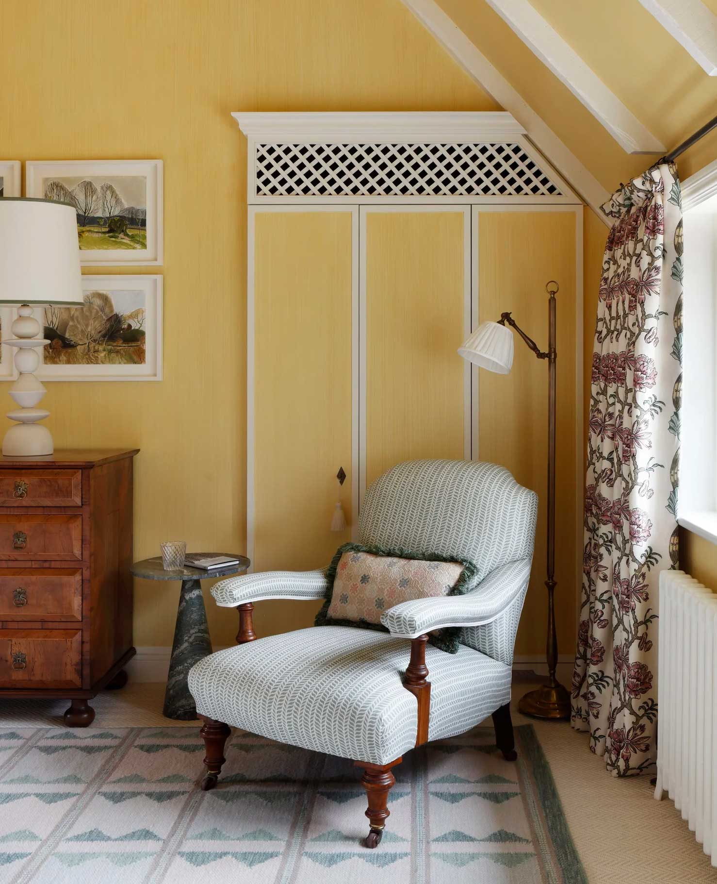 A cozy reading corner with a cushioned armchair, patterned pillow, wooden side table, standing lamp, floral curtains, a chest of drawers, framed art, and soft yellow walls with white trim. Natural light fills the space.