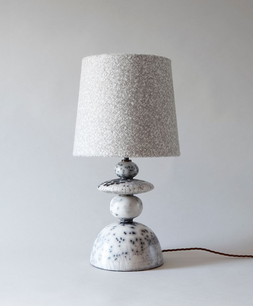 A modern table lamp with a textured, light gray fabric lampshade and a sculptural white marble base featuring black speckles, set against a plain light background. A brown cord extends from the base.