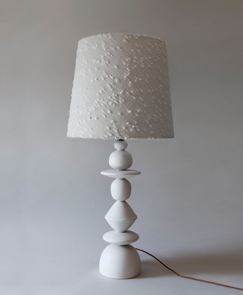 A modern table lamp with a textured white lampshade and a sculptural, geometric white base, set against a plain light gray background.