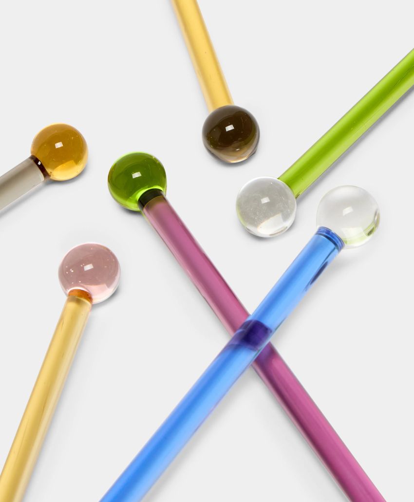 Six colorful glass stir sticks with rounded ends are arranged on a white surface. Each stick is a different color, including yellow, green, pink, blue, and clear.