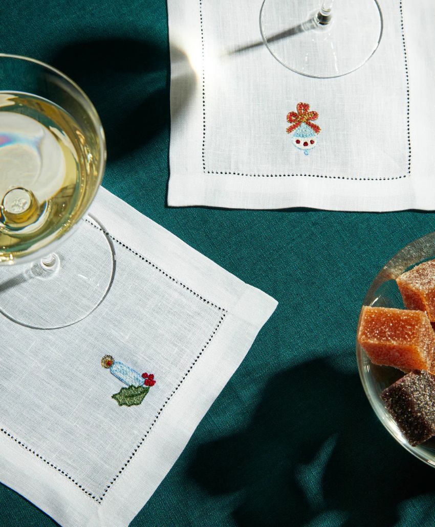 Two wine glasses sit on white embroidered cocktail napkins atop a teal tablecloth; one napkin has a snowman, the other an angel. A glass dish with assorted sugar-coated candies is nearby.