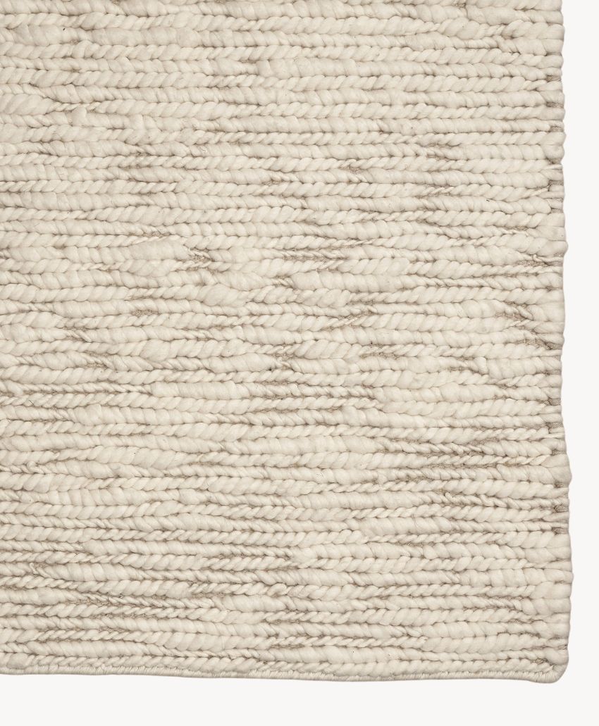 Close-up of a beige woven rug with a textured, chunky knit pattern and subtle variations in color. The edge of the rug is visible on the right side of the image.