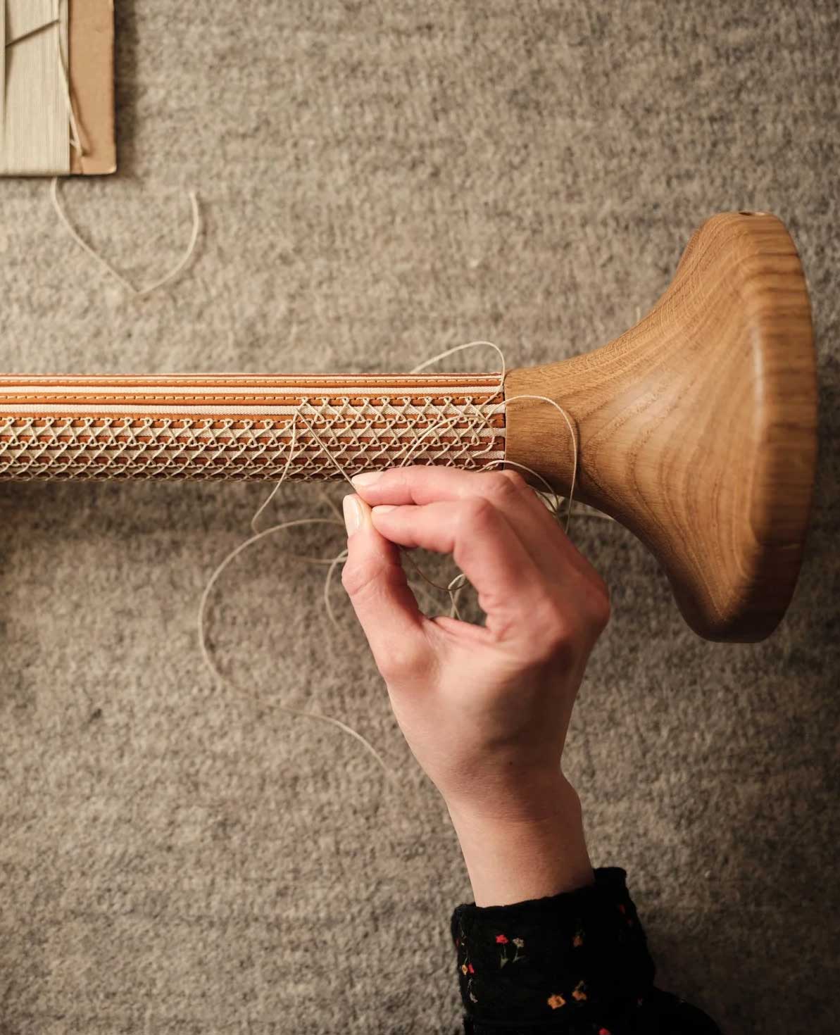 A hand is weaving or stitching thin thread through a wooden object with an intricate lattice pattern, possibly a furniture part, against a textured fabric background.