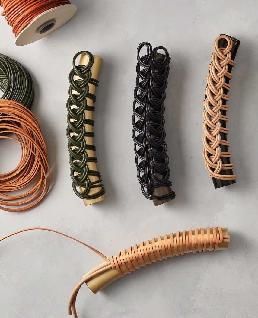 Four handles wrapped with intricate braided cord designs in green, black, and tan are displayed on a gray surface. Spools of matching cord are nearby, showing the materials used for the decorative handles.