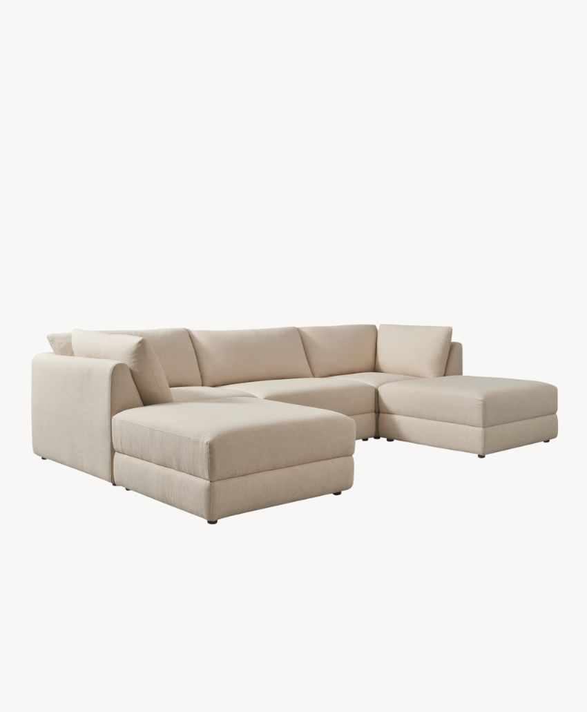 Beige sectional sofa with a chaise lounge on the left side and an extended seat on the right, featuring low, wide armrests and plush cushions against a plain white background.