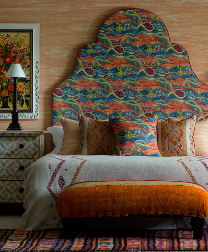 A colorful bedroom features a unique, patterned headboard, matching pillows, a white bedspread with designs, an orange bench at the foot of the bed, a nightstand with a lamp, and a framed floral painting on the wall.