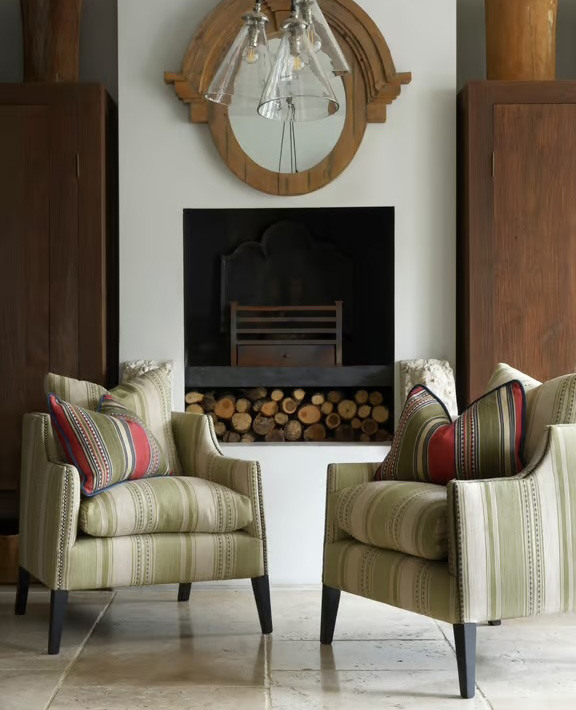 Two striped armchairs with red-accented pillows face each other in front of a fireplace filled with stacked firewood. Above the fireplace is a round wooden-framed mirror with pendant lights hanging down.
