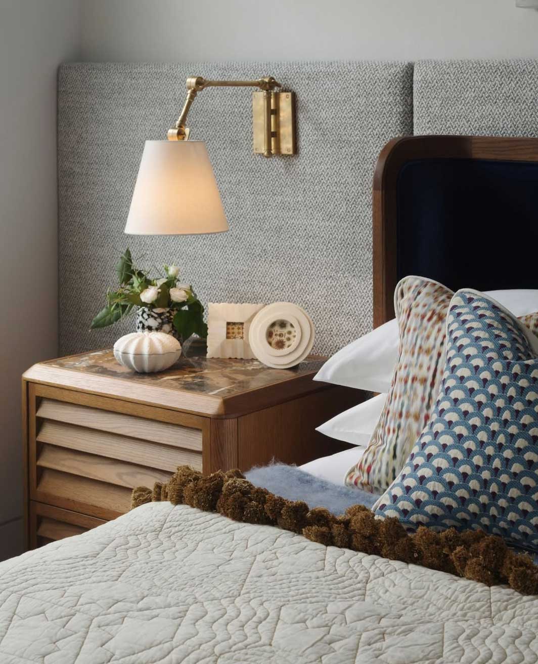 A cozy bedroom scene with a bed featuring patterned pillows and a textured quilt. A bedside table holds a small vase with flowers, a decorative white object, and a clock. A wall-mounted lamp provides light above the table.