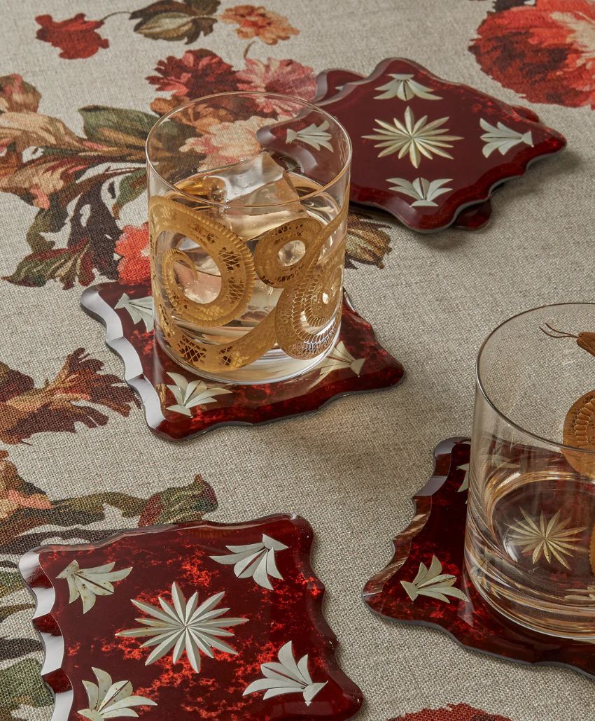 Two glasses with drinks sit on dark red coasters with white starburst designs, placed on a floral-patterned tablecloth. Additional coasters are nearby.