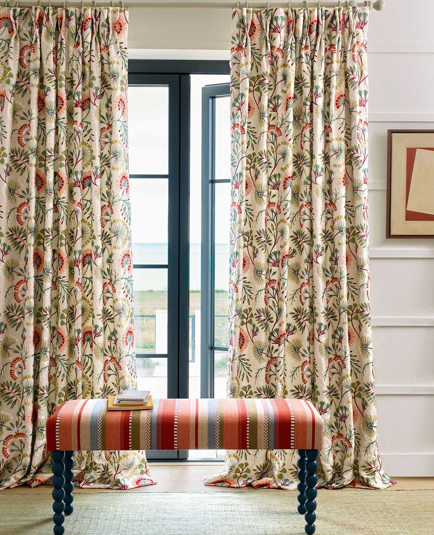 Two floral-patterned curtains hang on either side of a glass door. In front, a bench with red, orange, and white stripes sits on a green rug. A framed artwork hangs on the wall to the right.