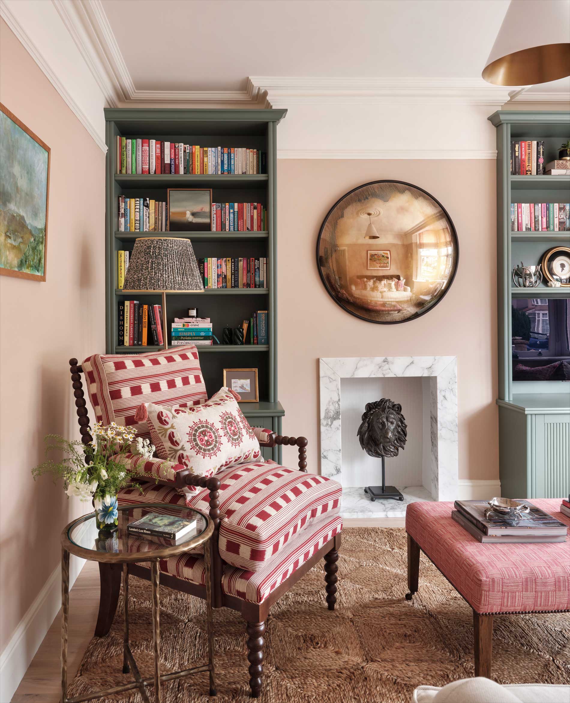 A cozy living room with patterned armchair and ottoman, glass side table, bookshelves, framed artwork, and a round mirror above a white fireplace with a black sculpture inside. Neutral walls and a textured rug complete the space.
