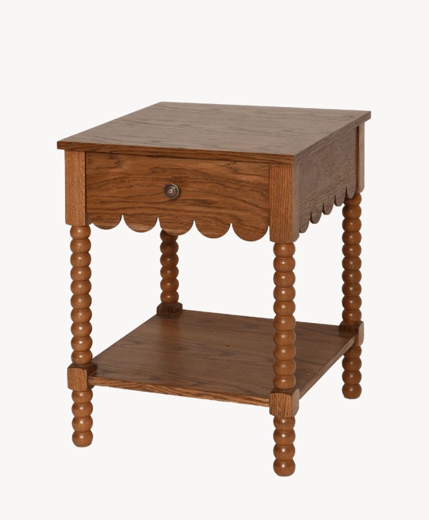 A wooden side table with turned legs, a single drawer featuring a round knob, scalloped detail beneath the drawer, and an open lower shelf.