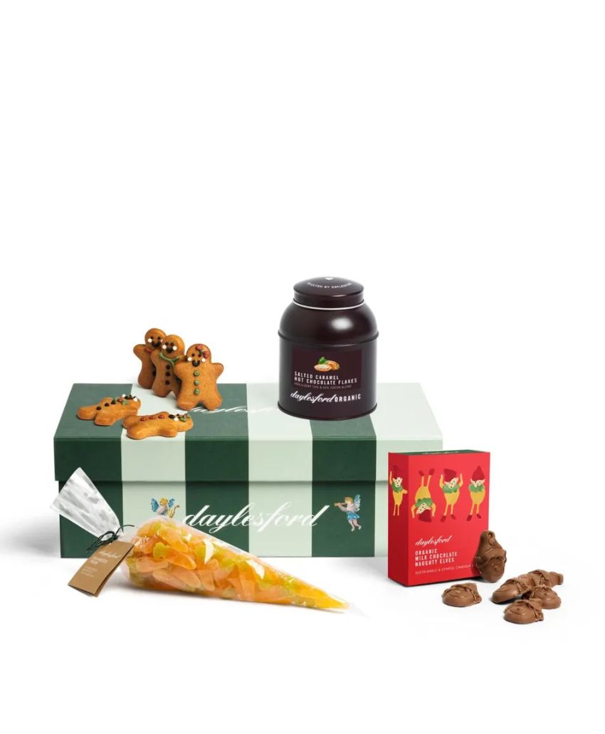 A gift box containing gingerbread cookies, a jar of spread, a piping bag with yellow filling, a red packet of chocolates, and chocolate pieces arranged on a white background.