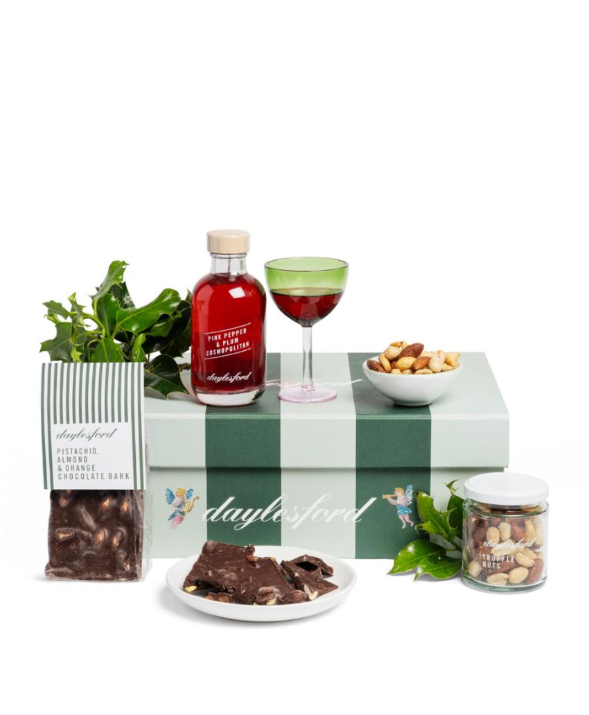 A green Daylesford gift box with a bottle and glass of red drink, a bowl of mixed nuts, a jar of treats, a bag of chocolate ginger cake, chocolate pieces on a plate, and green leaves arranged around.
