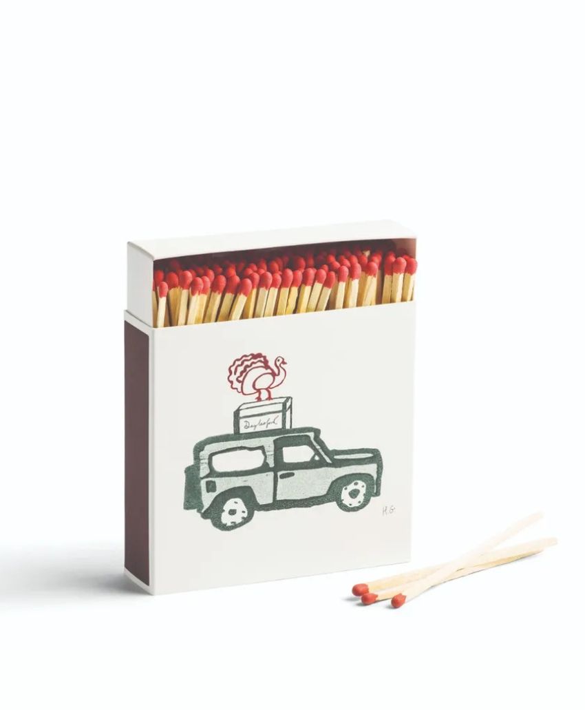 A box of matches with red tips, featuring an illustration of a vintage car carrying a rooster sign on its roof. Two loose matches lie next to the box on a white background.