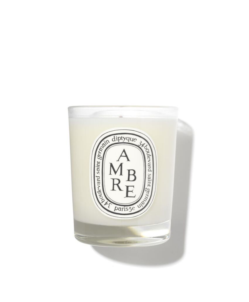 A white Diptyque scented candle in a clear glass jar with a white label that reads AMBRE and decorative black text around the edges, casting a soft shadow on a white background.