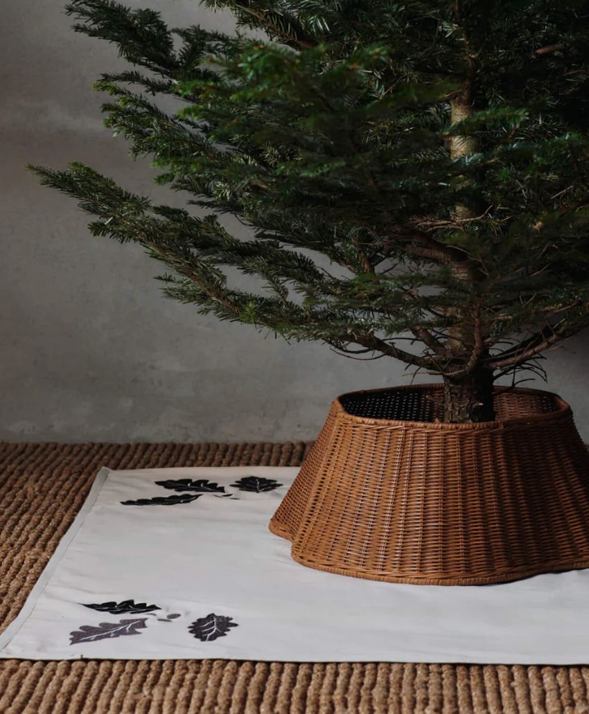 A pine tree in a wicker tree collar stands on a woven rug. The base is covered with a white cloth decorated with embroidered black leaves. The background is a plain, light-colored wall.
