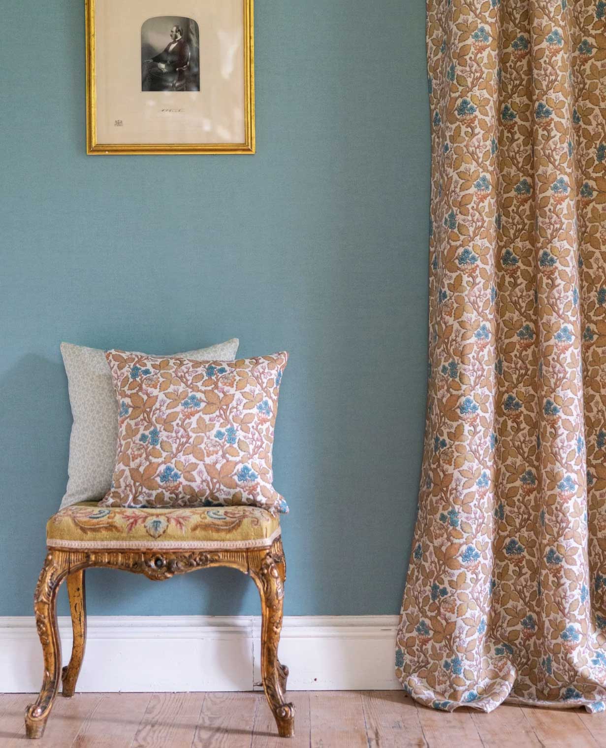 A decorative chair with a patterned cushion sits against a blue wall, next to matching floral curtains. Above the chair hangs a framed black-and-white portrait. The room has a wooden floor and white baseboards.
