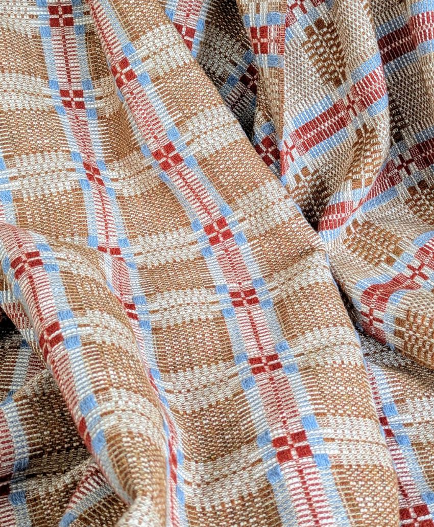 A close-up of woven fabric featuring a plaid pattern in beige, light blue, red, and white colors. The fabric appears soft and slightly draped with visible texture and intersecting colored lines.