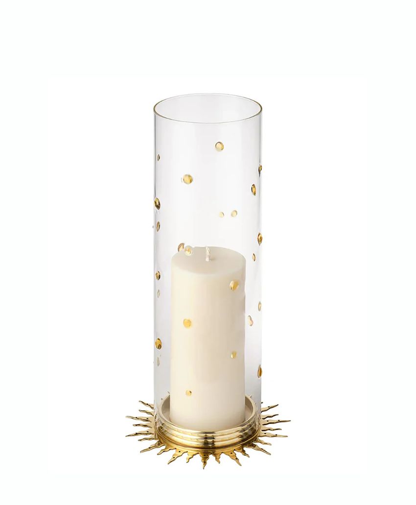 A white pillar candle sits inside a tall, clear glass hurricane decorated with small gold dots, placed on a round gold base with sunburst-like rays extending outward.