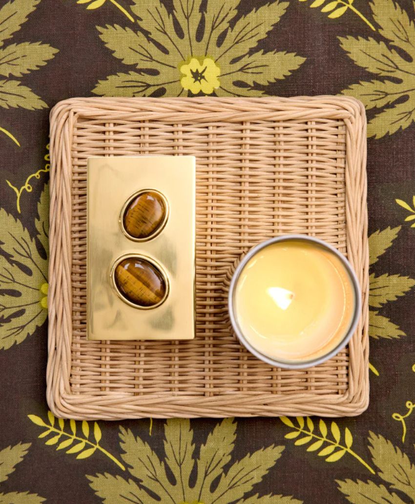 A gold box with two oval stones and a lit yellow candle sit on a woven tray, placed on a brown and green leaf-patterned fabric background.