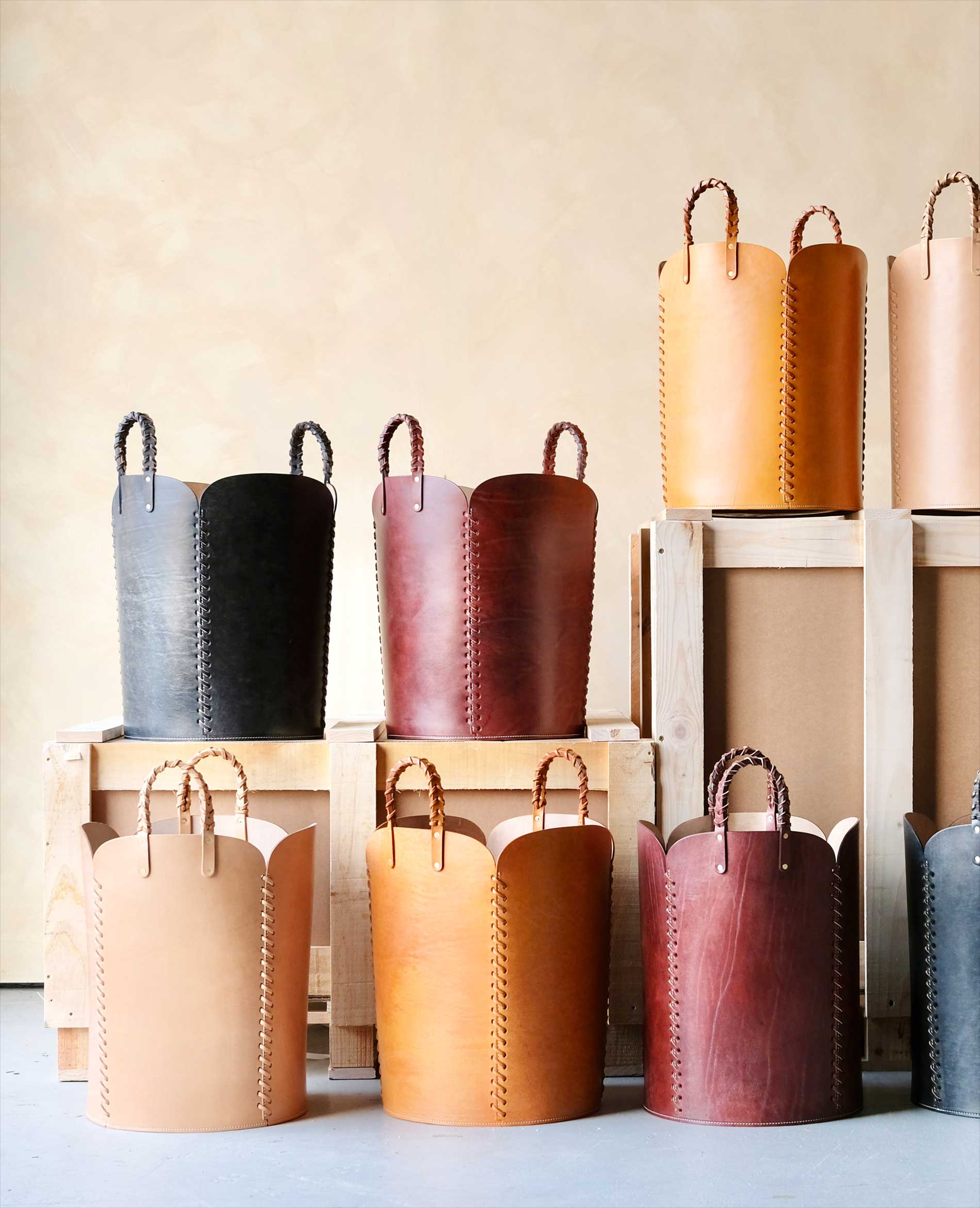 Several tall, leather baskets with braided handles in various shades of brown, tan, and black are displayed on and around wooden crates against a neutral background.